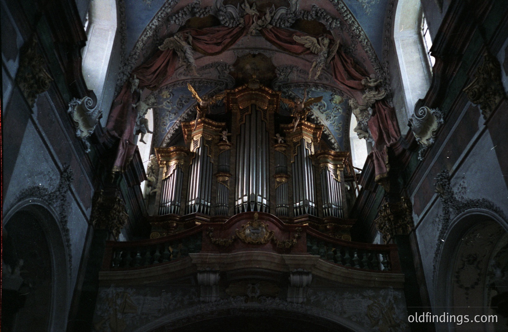 Ornate, gilded pipe organ dominates a baroque church interior. Elaborate carvings & painted frescoes featuring cherubic figures embellish the surrounding architecture. Likely late 17th or early 18th century European construction. Fine example of period craftsmanship.