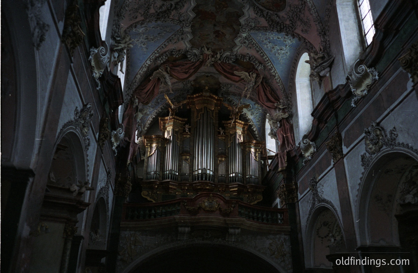 Ornate Baroque organ dominates a church interior. Elaborate carvings, dark wood, and painted ceiling with cherubic figures create a dramatic effect. Arched niches frame the view, emphasizing the building's scale and grandeur. Likely 18th-century European design.