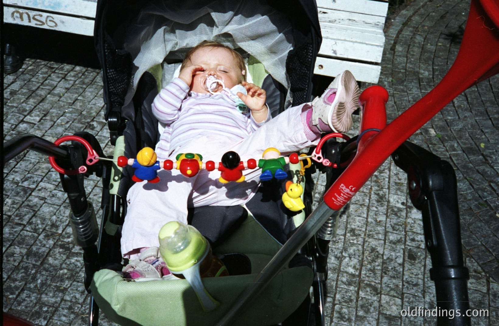 Infant asleep in a green stroller, pacifier visible. Red stroller frame, baby toys dangling above. Likely a candid, casual snapshot. Commercial potential for parenting/baby product stock. Possibly 1990s-2000s, given stroller style.