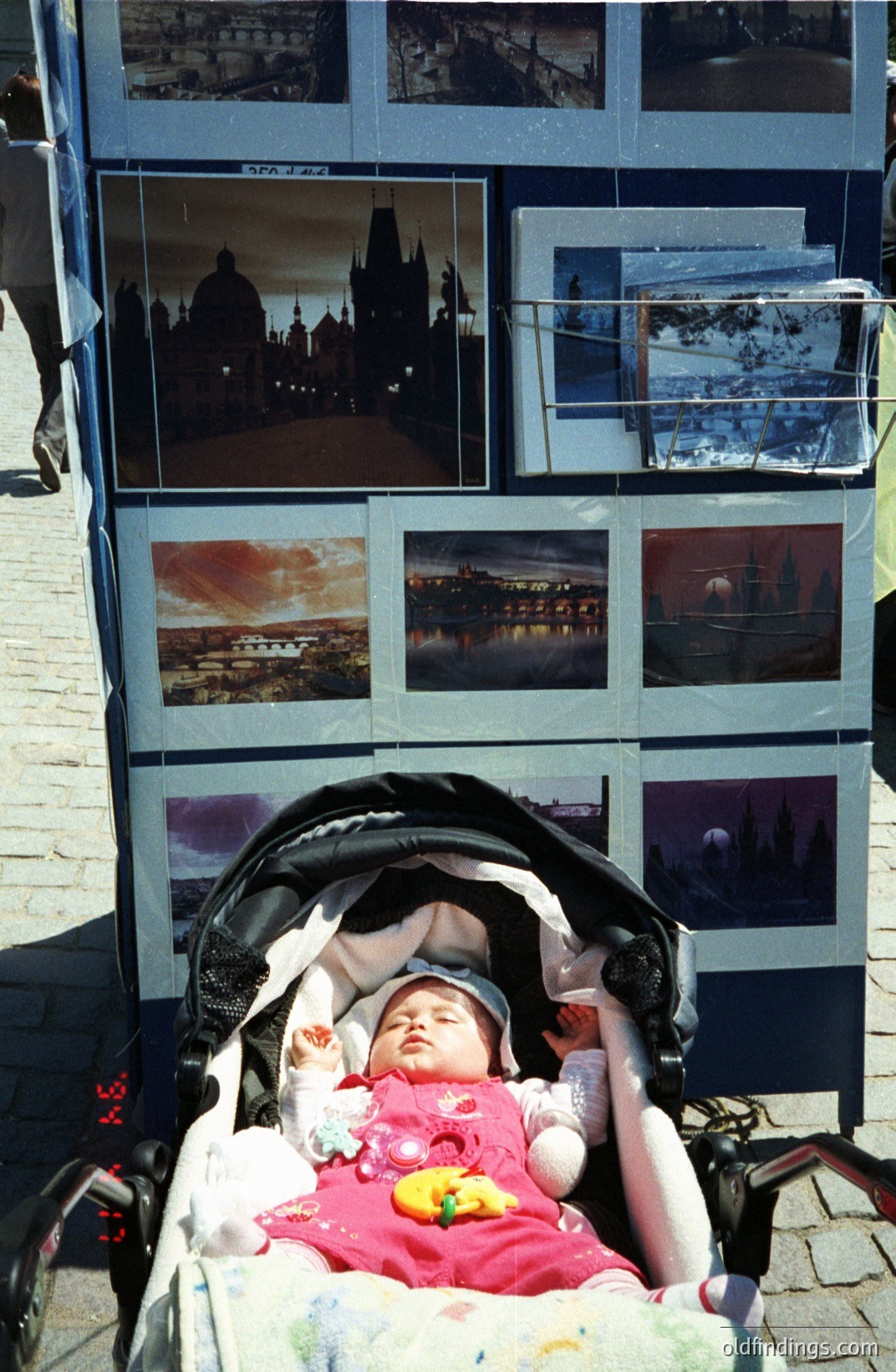 A baby sleeps peacefully in a stroller, swaddled in pink and white layers. The backdrop is a display of landscape photographs, likely for sale. Appears to be a tourist area, possibly Europe, 1990s-early 2000s based on film style & stroller design. Commercial value: stock photo reference for travel/childhood.