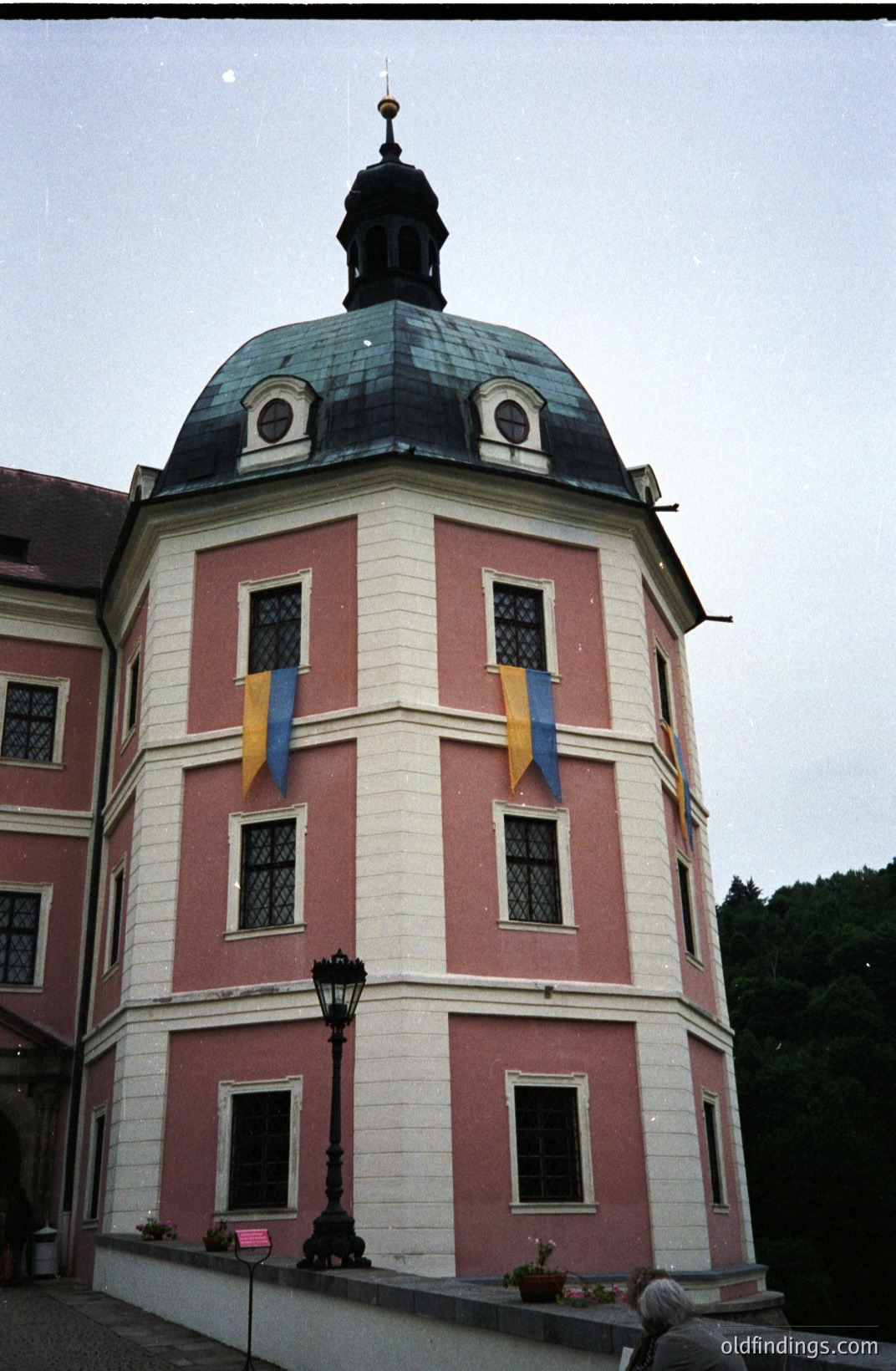 Elaborate, pink and white stucco architecture with a distinctive domed roof. A formal garden with a lamppost and plantings is visible in the foreground. Flags hang from the building's facade. Likely a stately home or castle.