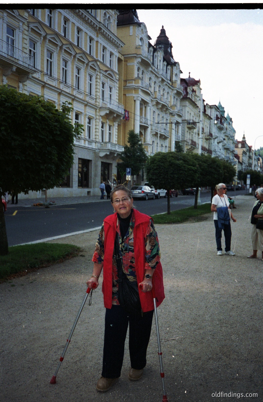 An older woman stands using walking sticks before a grand, ornate building. She wears a red jacket over a patterned top and dark pants. Lush, manicured trees line a wide street. Architectural style suggests Central/Eastern Europe, likely early 20th century. Likely a tourist destination.