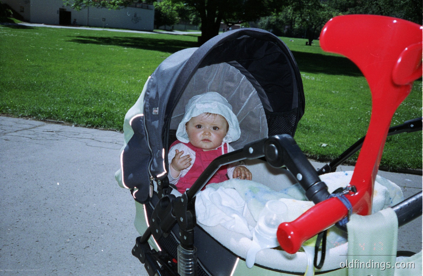 Infant in a stroller, shaded by a fabric canopy. The child wears a white bonnet. The stroller features a bright red handlebar. Appears to be a candid, possibly amateur photograph. Likely 1980s or 1990s based on the stroller’s design.