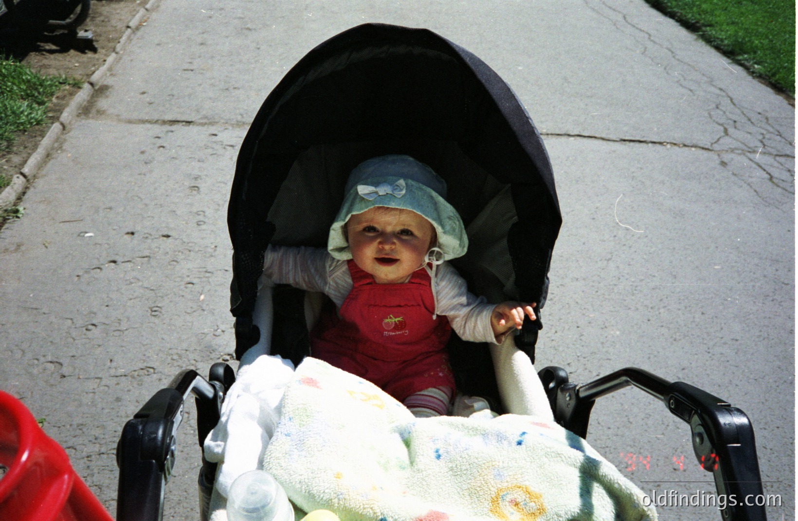 Infant sits in a vintage stroller, wearing a patterned hat and red overalls. A light-colored blanket peeks from beneath. The scene suggests a casual, everyday moment in the 1970s or early 1980s. Commercial value: stock photo reference for family or childhood themes.