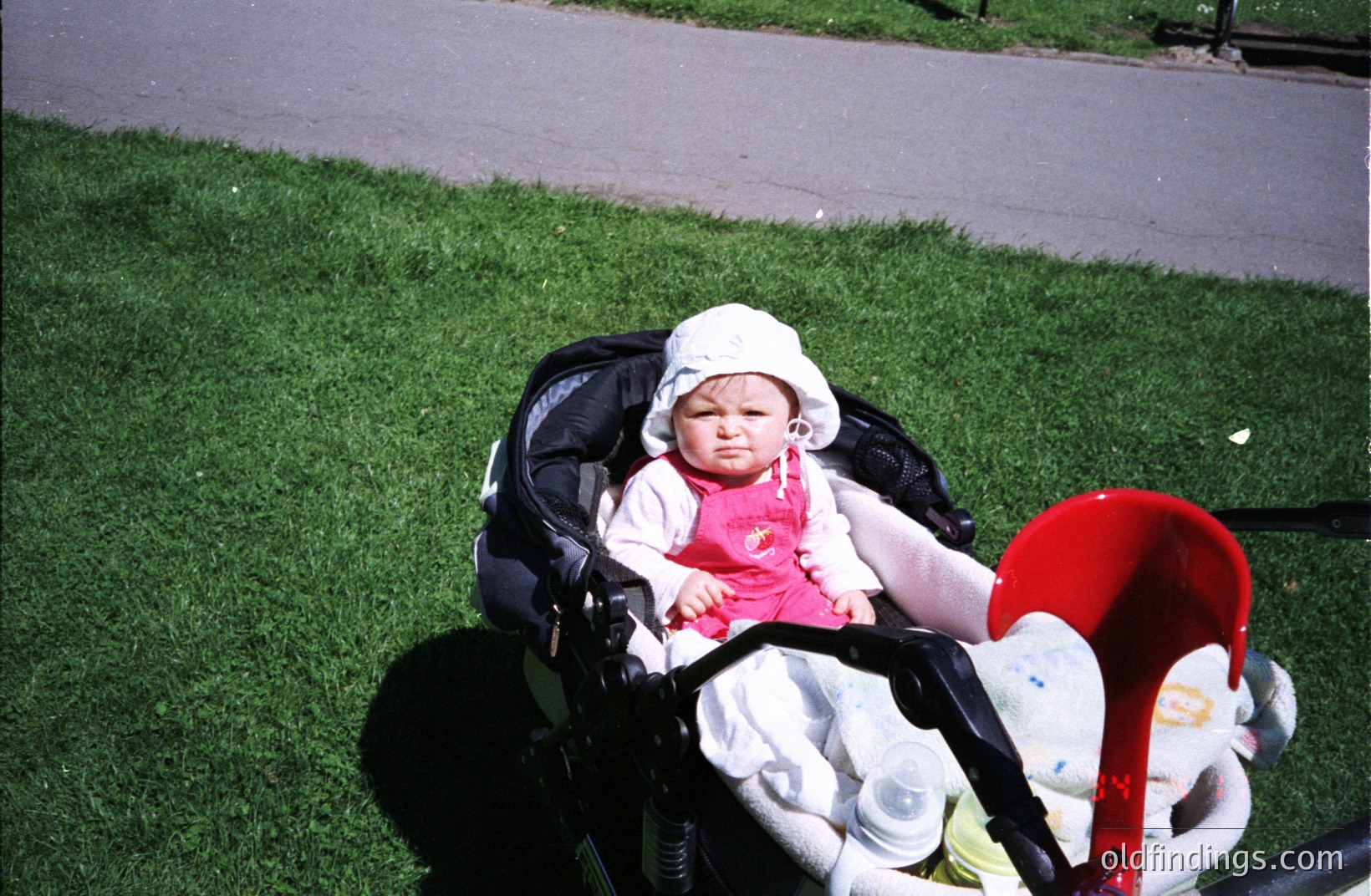Infant seated in a baby stroller on green grass, wearing a pink dress and white hat. The stroller features a red safety bar and a plush toy. Likely a candid, casual moment, possibly a family outing.