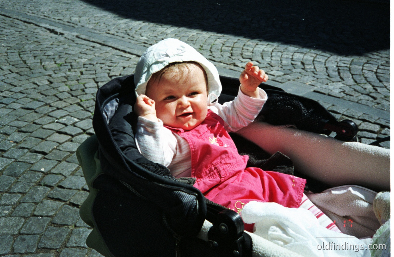A smiling infant sits in a stroller, dressed in a pink dress and white bonnet. The background shows a stone-paved surface. Likely a candid, casual portrait. The image appears to be from the 1980s-1990s based on the stroller design.