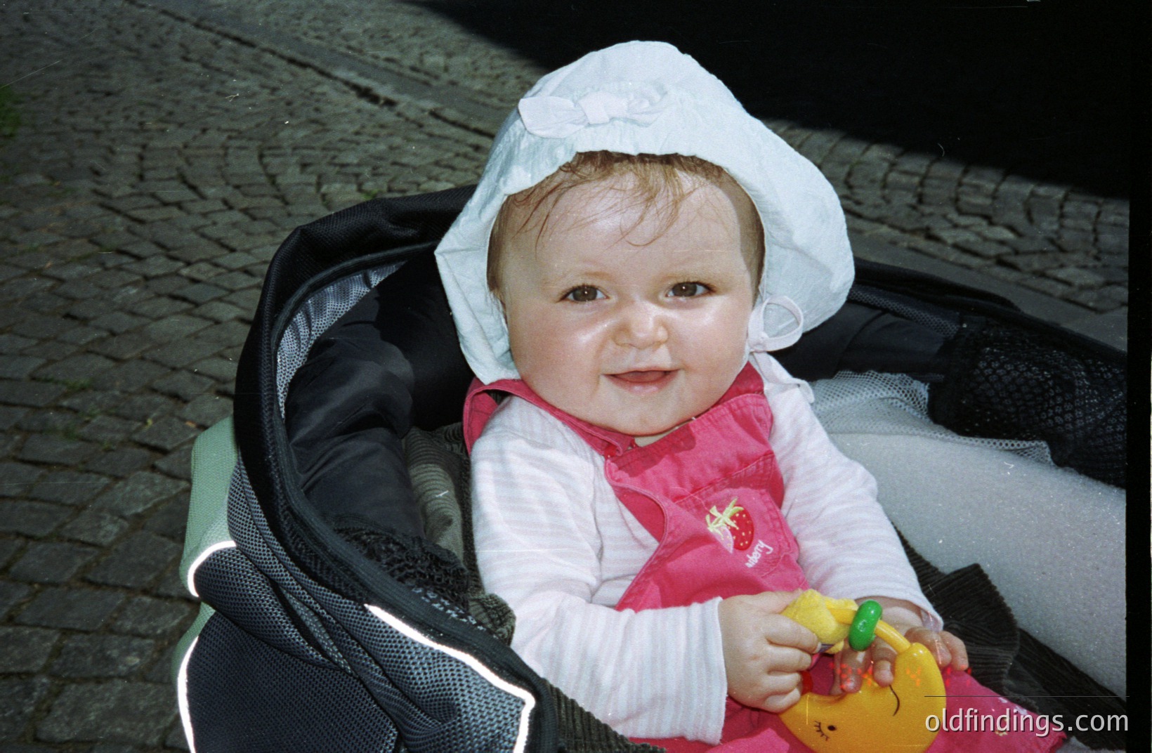 Infant in a stroller, wearing a white bonnet and pink dress. Holding a rubber duck toy. Likely a family snapshot, potentially from the 1990s or early 2000s. Background features cobblestone pavement. Stock potential for baby/childhood themes.