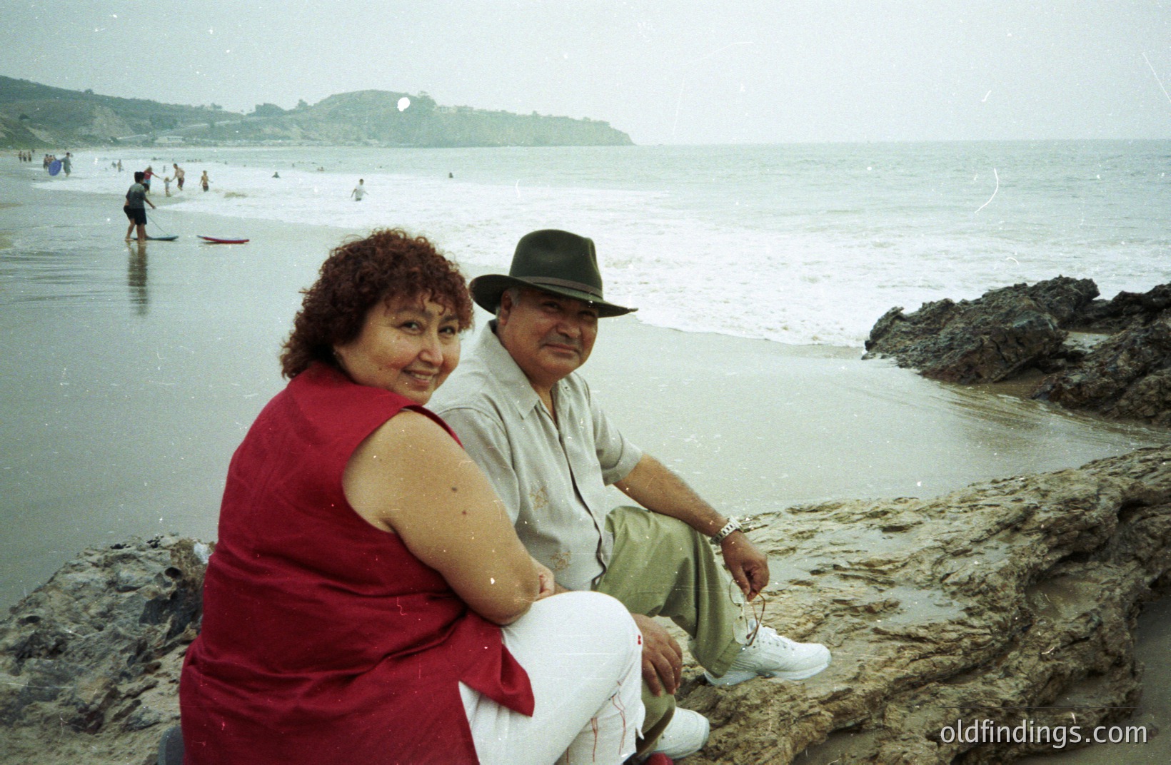 A couple sits on rocky coastline overlooking a sandy beach. The woman wears a red tank top; the man a fedora and white pants. Several figures are visible wading in the water. Likely a vacation photo, potentially from the 1970s or 80s based on clothing styles.