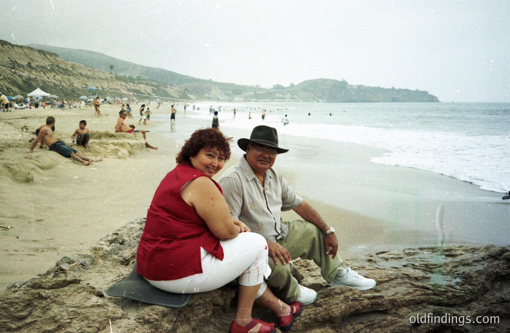 A couple sits on a rocky outcrop overlooking a sandy beach with waves gently rolling in. The man wears a fedora and light-colored pants; the woman, a red top and white pants. Background shows a crowded beach and sloping hills. Likely Southern California, 1980s-90s.