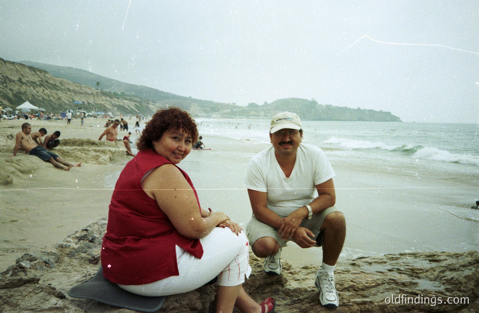 A couple sits on rocks overlooking a sandy beach. The woman wears a red top and white capri pants. The man is in a white t-shirt and khaki shorts, sporting a baseball cap. A rocky coastline & distant beachgoers are visible. Likely 1990s casual snapshot.