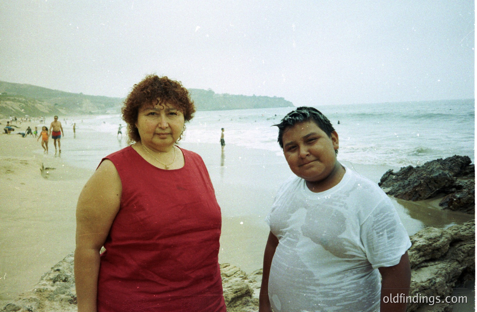 A woman and boy stand on a sandy beach, likely a family vacation snapshot. The woman wears a red sleeveless dress; the boy, a wet, clinging white t-shirt. Background features ocean waves, rocky cliffs, and other beachgoers. Likely 1980s-1990s amateur photography.