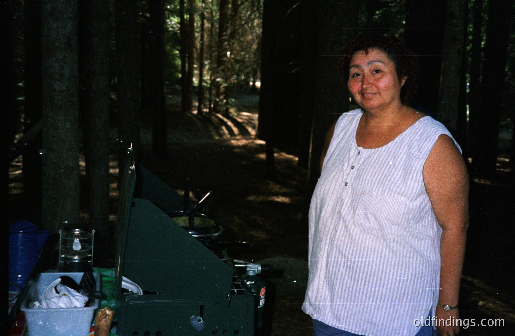 A smiling woman stands outdoors beside a camping stove and equipment, possibly for cooking. Forested background with filtered sunlight. She wears a striped sleeveless shirt and a watch. Appears to be a casual, candid snapshot. Likely 1980s or 1990s home photography.