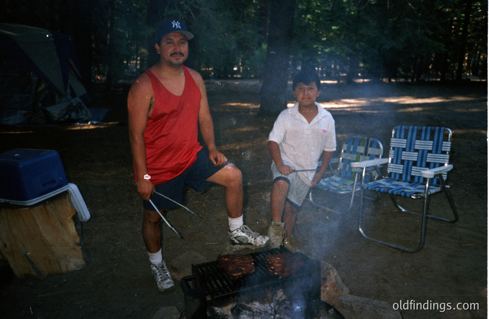 A man and boy grill food over a campfire. The adult wears a red tank top and baseball cap, while the boy is in a short-sleeved shirt. A cooler and firewood are nearby. Likely a family camping trip, possibly in the 1980s or 1990s based on clothing styles.
