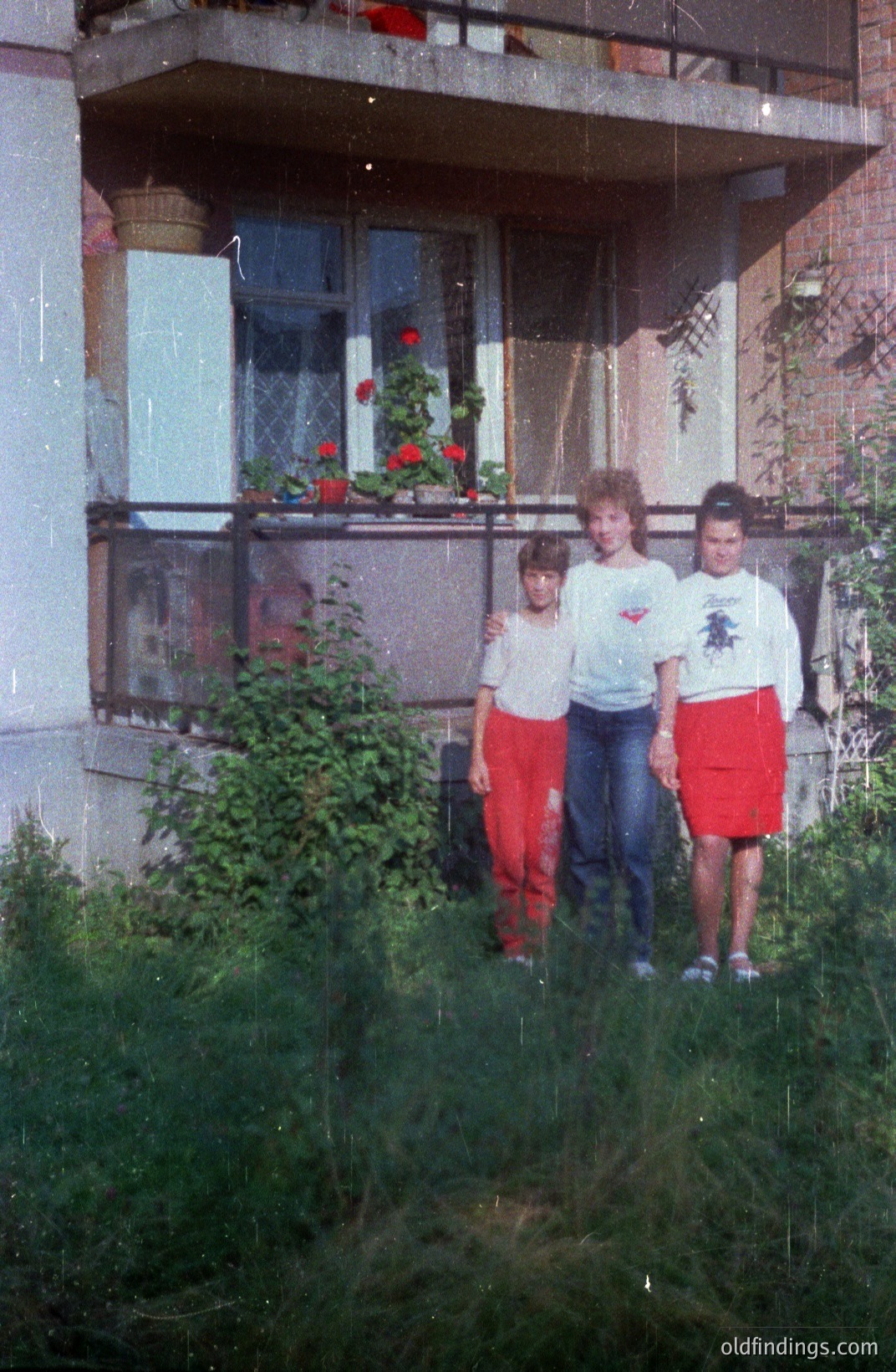 Three children stand in overgrown grass before a block of flats. The building features a balcony adorned with potted geraniums. Clothing suggests a casual, late 1970s/early 1980s style. Likely Eastern European location, potentially a socialist housing project. Strong composition, archival value. flats