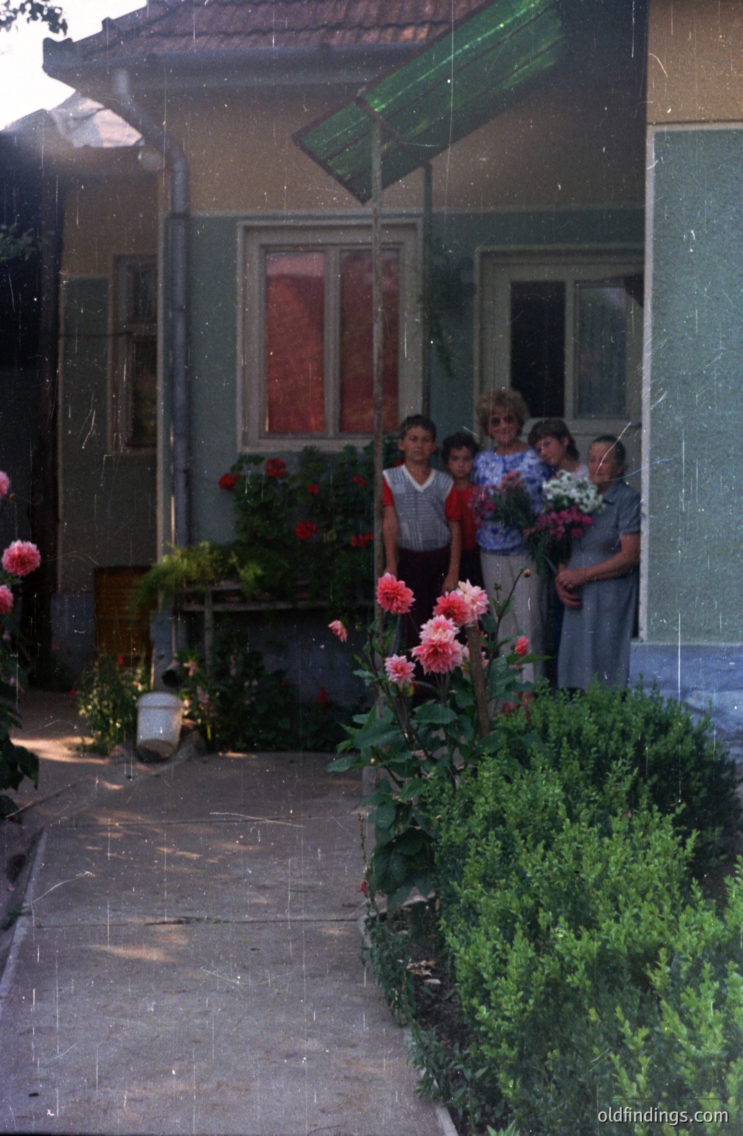 A group of four young people stands in a doorway, holding bouquets of flowers. The scene appears to be a residential courtyard with a simple, pale green building and lush greenery. Likely Bulgaria, 1970s-80s judging by the style and film quality. A moped is visible in the background.