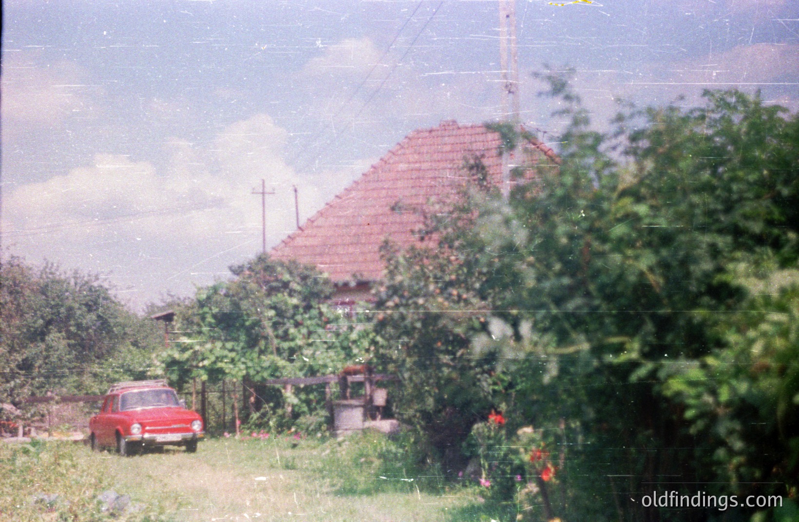A red Lada car sits on a rural road with a traditional, terracotta-roofed house partially obscured by dense foliage. Likely Eastern Europe, circa 1970s-80s, based on vehicle and architecture. Rustic scene, indicative of everyday life in a socialist country. Offers unique aesthetic for design or archival purposes.
