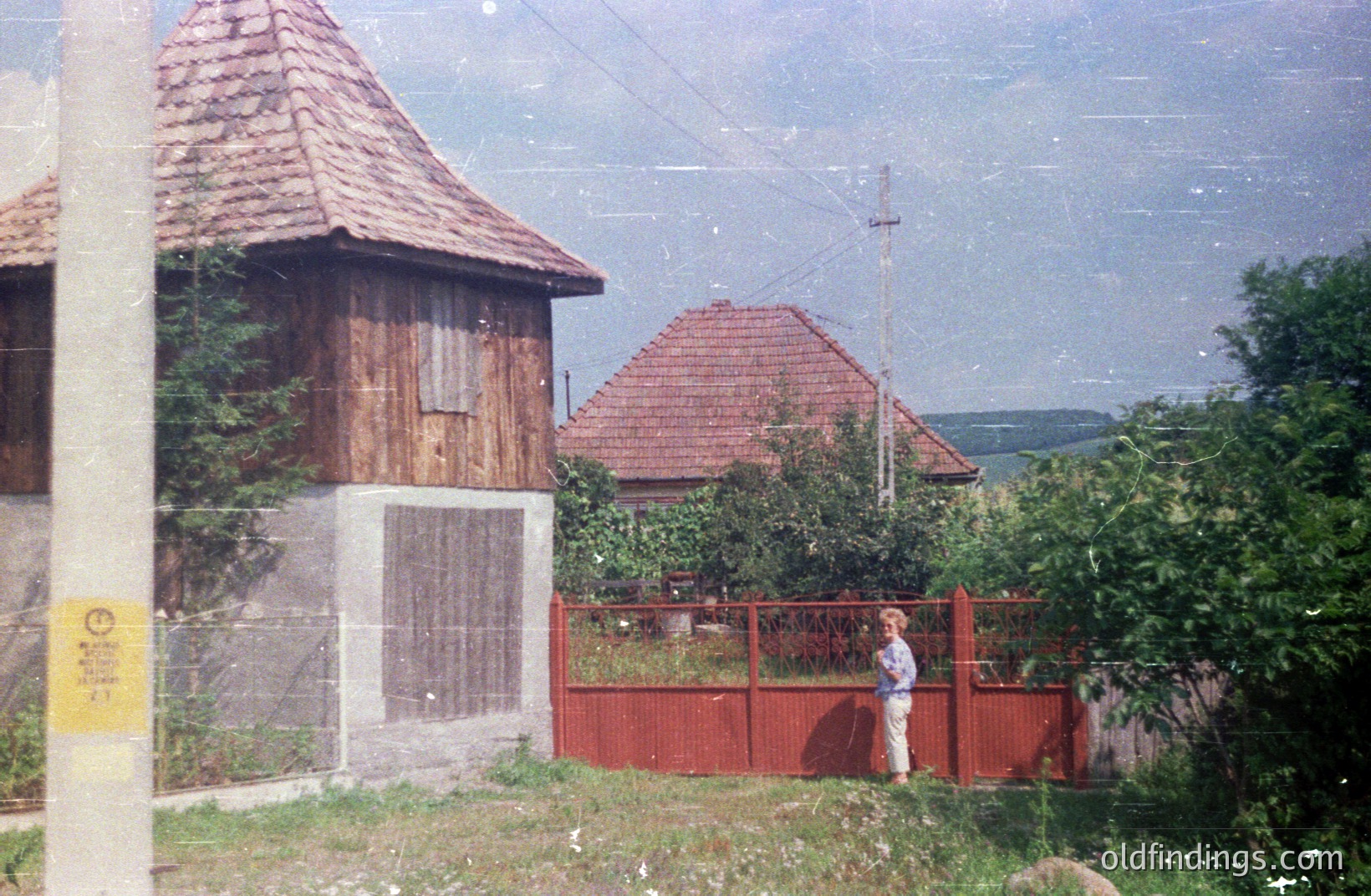 A weathered, tiled-roof building stands adjacent to a red-painted picket fence and a figure stands beyond. The architecture suggests rural Eastern European design, potentially late 20th century. The view extends to a green, rolling landscape. Likely candid moment captured on film.