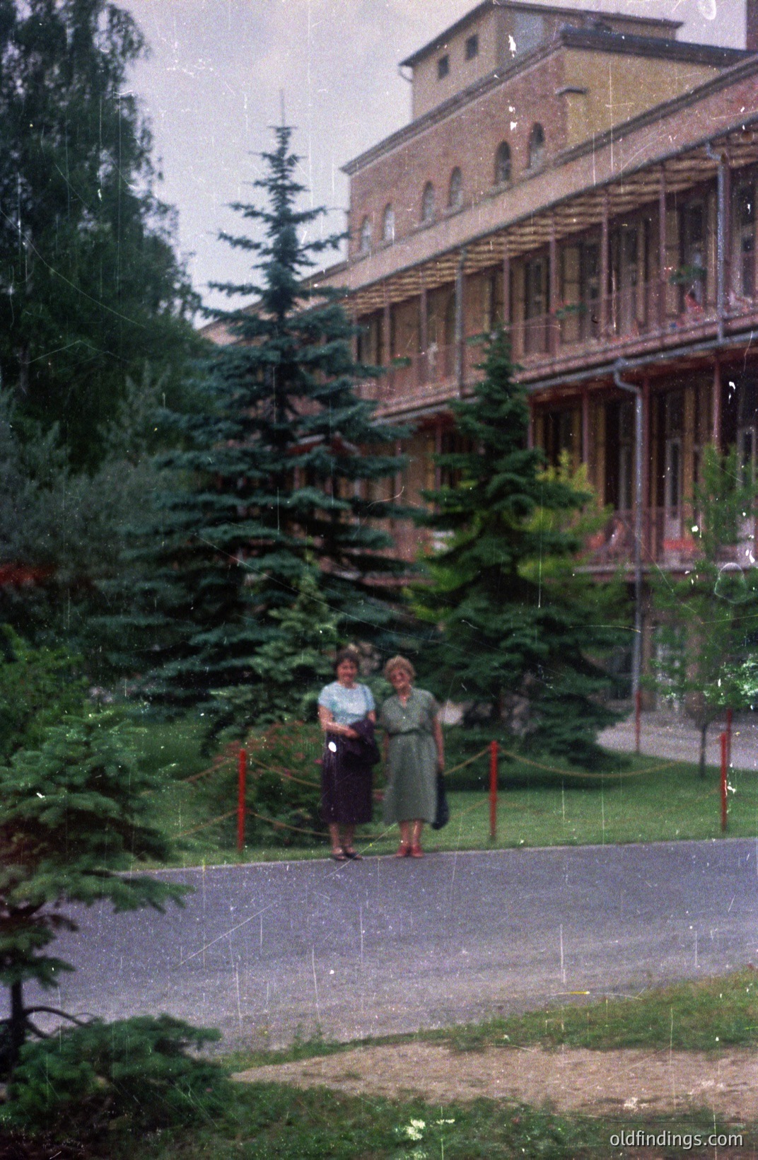 A 1970s color photograph depicts two women in knee-length dresses walking along a paved path alongside a grand, Soviet-era sanatorium building with a prominent balcony. Appears to be a seaside resort. Evergreen trees line the path. Good for design referencing.