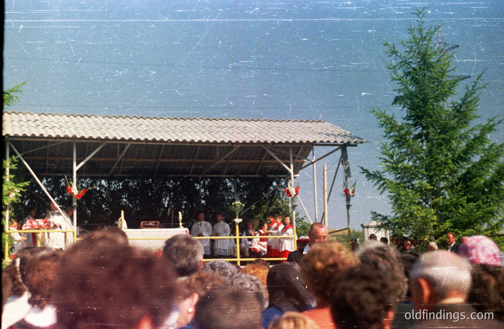 A partially visible crowd gathers beneath a metal-roofed platform. Individuals in white robes stand on stage facing a backdrop decorated with red elements. Seaside location suggested by a distant ocean view. Likely a ceremonial or religious event. Photographic quality suggests a snapshot from the 1970s or 1980s.