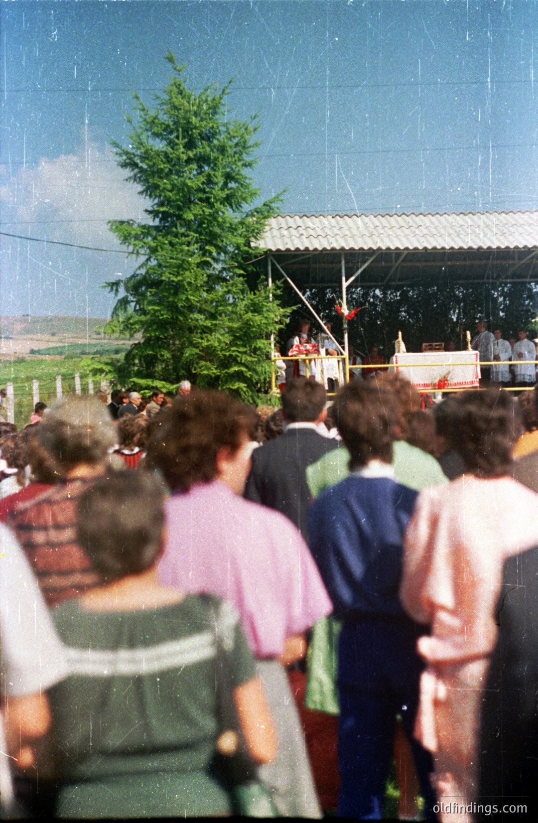 A crowd gathers outdoors before a makeshift stage with a religious ceremony underway. Attendees are seen from the rear, wearing 1970s-style clothing, indicating a likely timeframe. A tree stands prominently beside the structure. The backdrop features a field and distant hills.