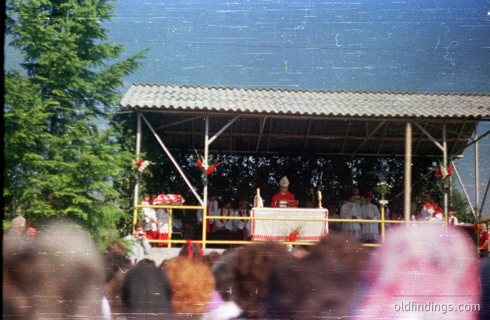 A religious ceremony unfolds beneath a simple, open-air pavilion, likely in a rural setting. Figures in vestments lead the service at a decorated altar. Crowd visible in the foreground suggests a public or community event. Appears to be a 1970s or 80s snapshot.