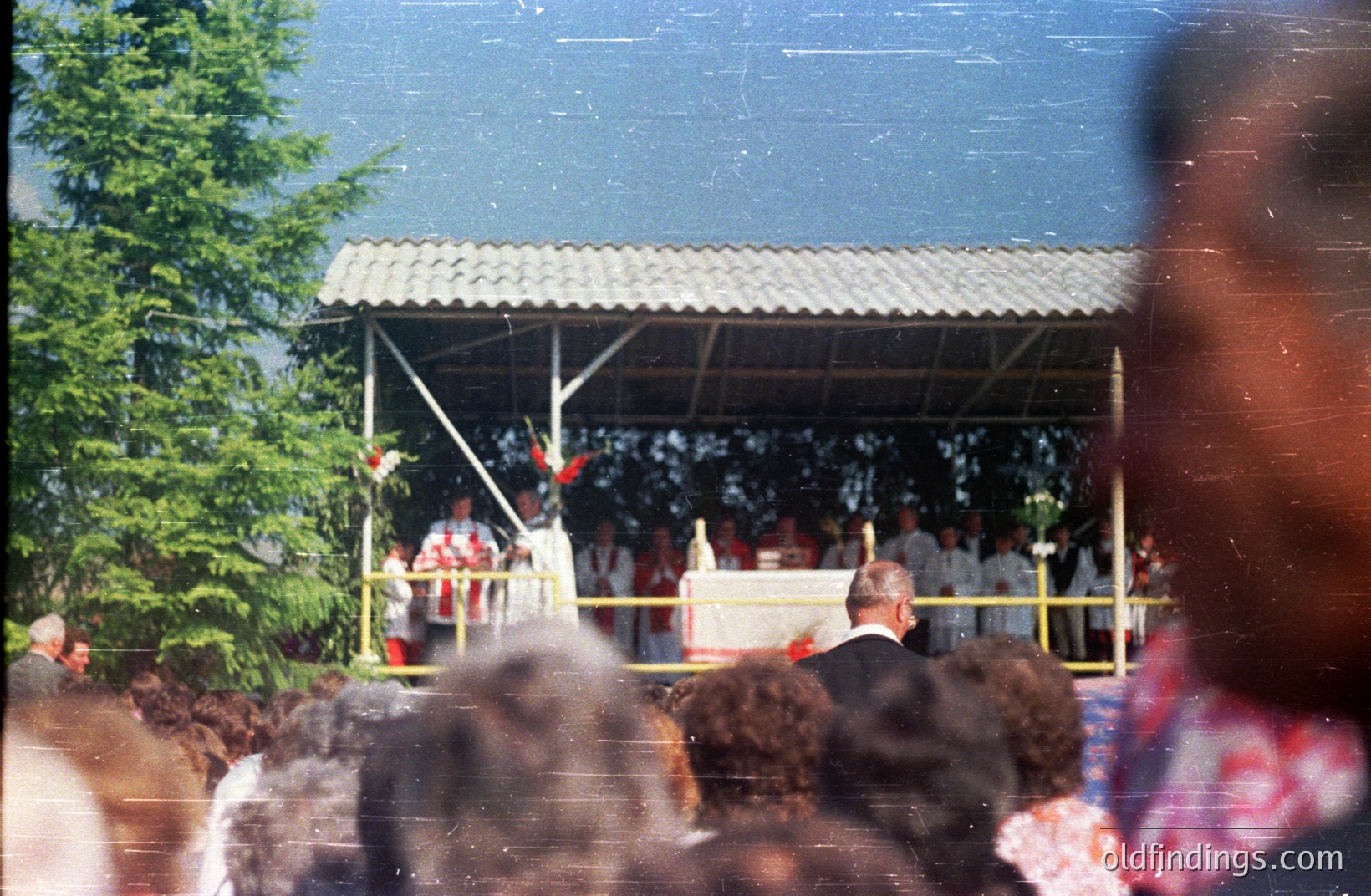Outdoor religious ceremony captured in a candid moment. A large crowd gathers under a simple, corrugated metal structure. A priest officiates at an altar adorned with flowers, likely a wedding or confirmation. 1970s color film aesthetic. Possible coastal location.