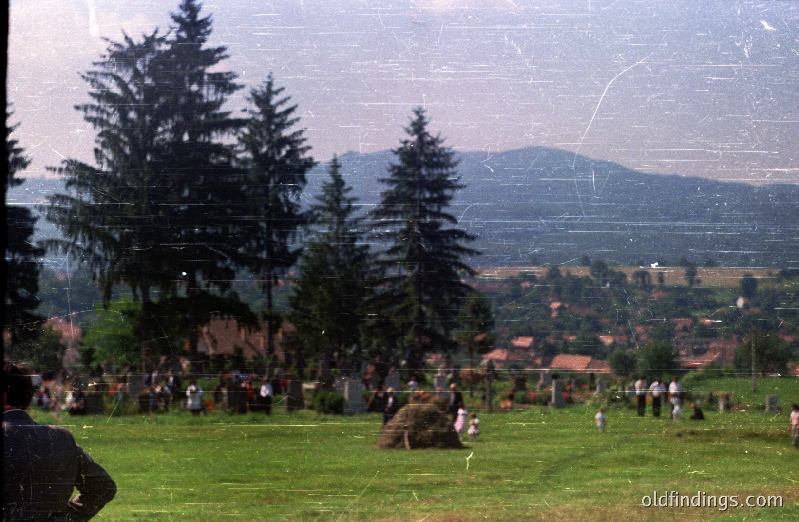 A hillside meadow hosts a gathering, likely a festival or public event. Traditional dress is visible among the attendees. Background reveals rural village houses & distant, forested mountains. Appears to be Eastern Europe, possibly Bulgaria. Film grain & color saturation suggest a 1970s timeframe.