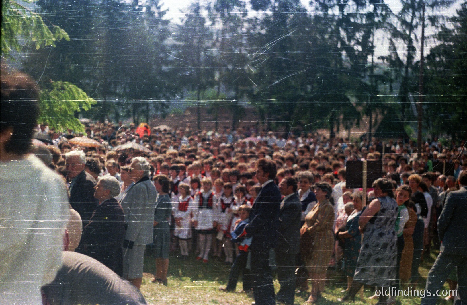 Large outdoor gathering with crowds of people, some in formal attire. Visible are children wearing white shirts and red neckties, likely a school group. Lush greenery and trees form a backdrop. Grainy image suggests a 1970s or 80s amateur photograph. Potential for historical documentation or design reference.