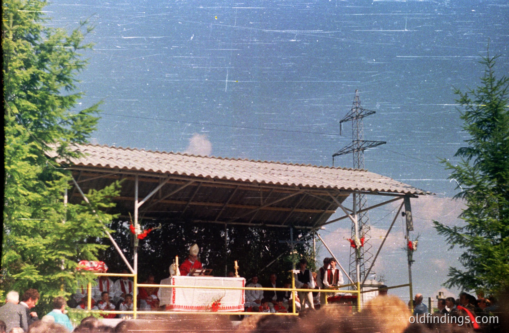 A wooden stage with a corrugated metal roof stands amidst lush greenery, likely an outdoor gathering space. Figures are visible on the stage and in the audience. Power lines dominate the background sky. Appears to be a public or ceremonial event.
