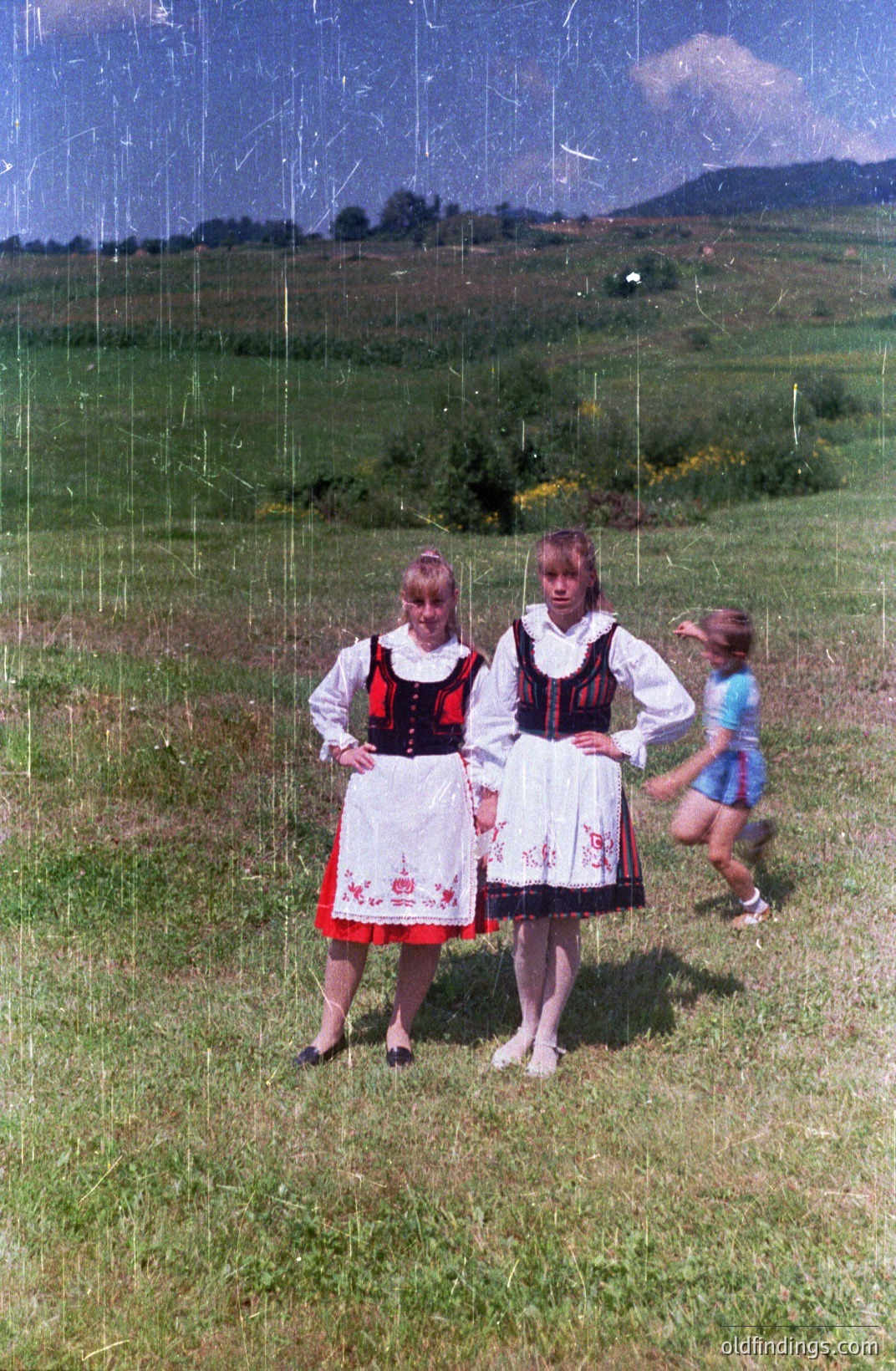 Two young women in traditional folk dress stand facing the camera amidst a grassy hillside. Detailed black & red embroidered bodices & white skirts are notable. A boy runs away from the scene in a blue t-shirt. Appears to be a rural, possibly Eastern European, setting. Likely 1970s.