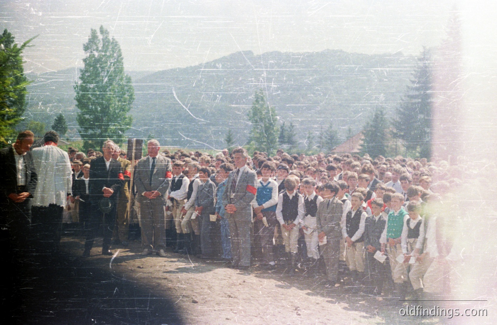 A large group of formally dressed boys stand in a line outdoors, likely schoolchildren. Men in suits observe the group, some with red pins. Background features a hilly, forested landscape, suggesting a rural location. Appears to be a staged or ceremonial event, likely 1960s-1970s Eastern Europe. Faded, with film grain and scratches.
