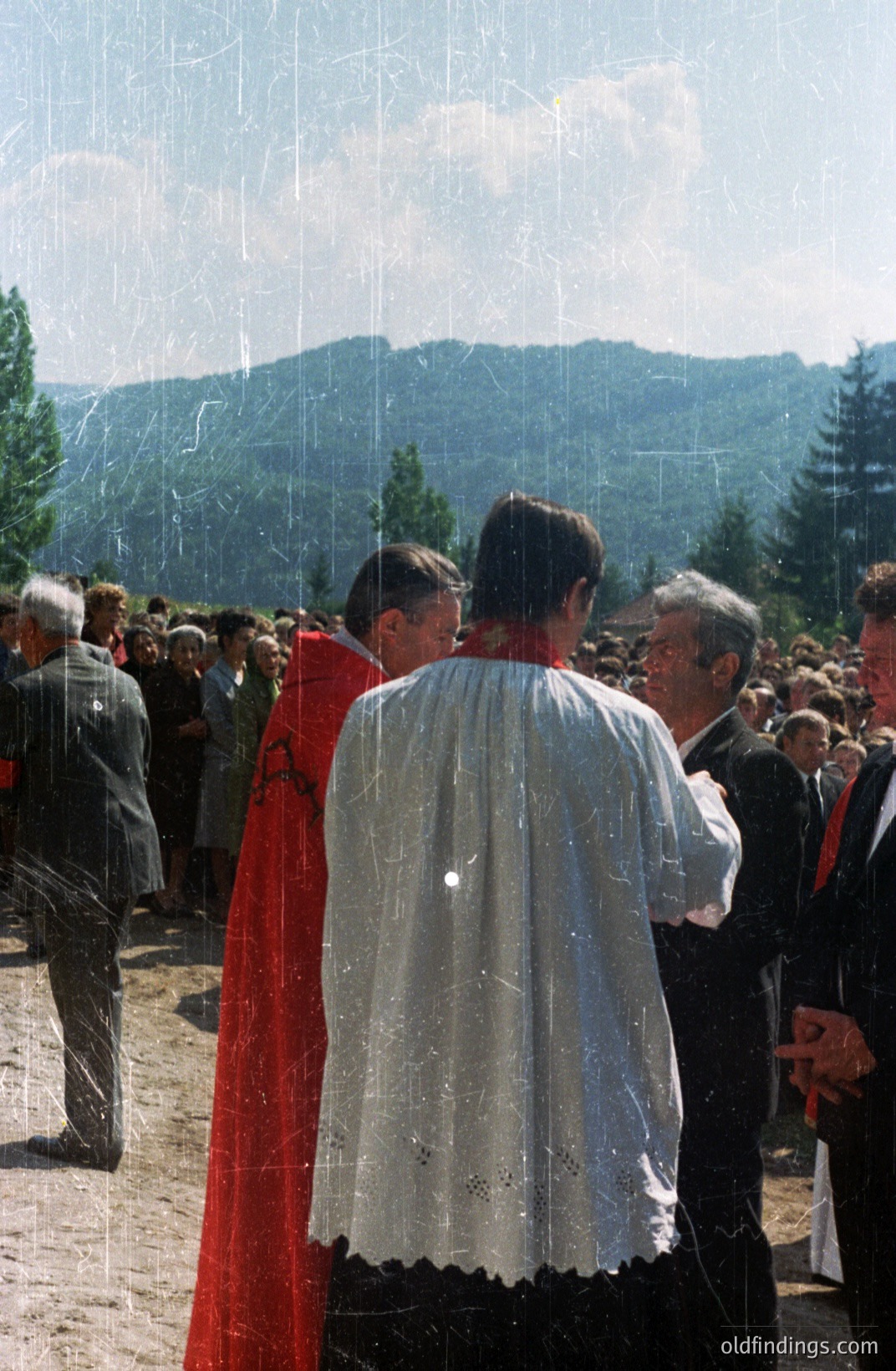 Formal gathering outdoors, featuring two figures in ecclesiastical vestments. One wears a white, intricately patterned robe; the other, a red cloak. A crowd of people is visible in the background, set against a forested, mountainous backdrop. Likely a religious ceremony, possibly in a rural European location. Appears to be a 1970s or 80s image based on film grain & clothing styles.