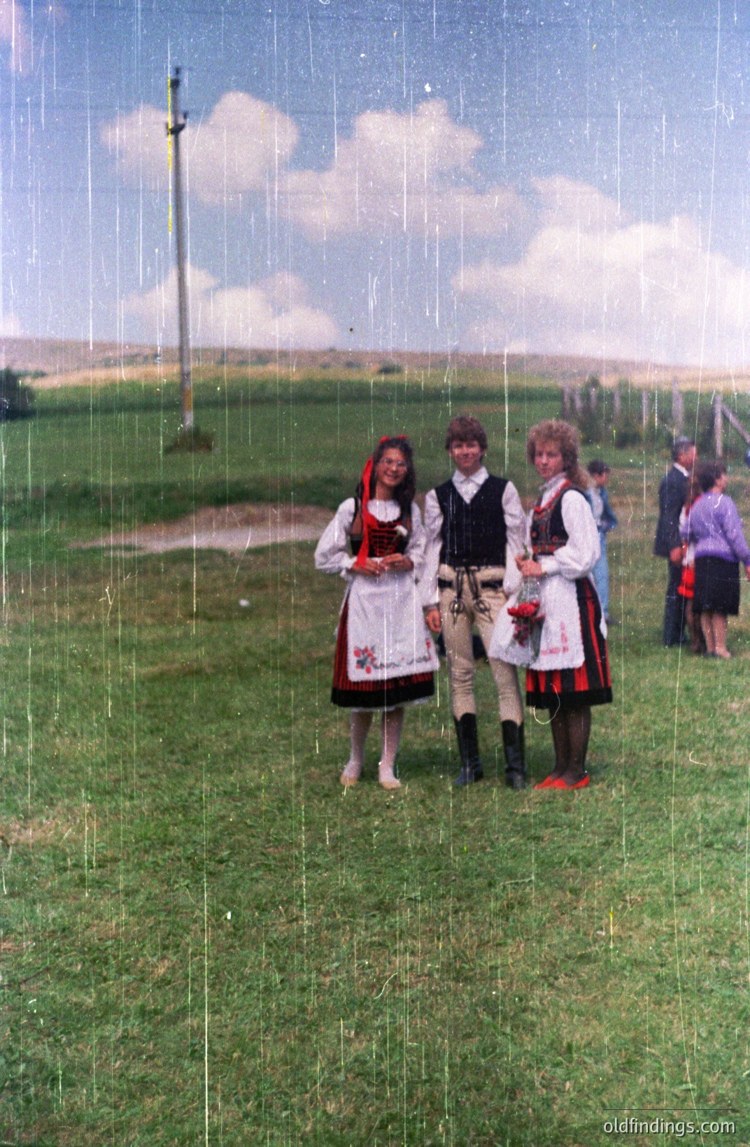 Three young people in traditional folk dress pose for a portrait on a grassy field. The scene includes lush green landscape, a rural setting with a distant treeline, and a power line. Likely Bulgaria, based on the folk costumes. Dating from the 1970s or 1980s, judging by the photographic style and clothing.