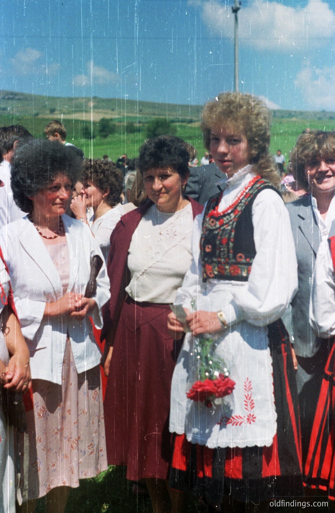Three women in traditional folk attire stand in a field alongside others at an outdoor gathering. The woman in the center wears a cream-colored blouse and maroon skirt, while the woman on the right is in a white embroidered dress and red sash. Photograph displays typical 1970s color palette.
