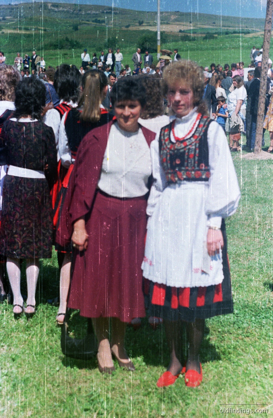 Two women stand in a field of green grass, both wearing traditional folk dress. The woman on the left wears a red skirt and jacket, while the other has a white blouse with red embroidery and a black skirt with red accents. A large crowd is visible in the background. Appears to be a festival or cultural event.