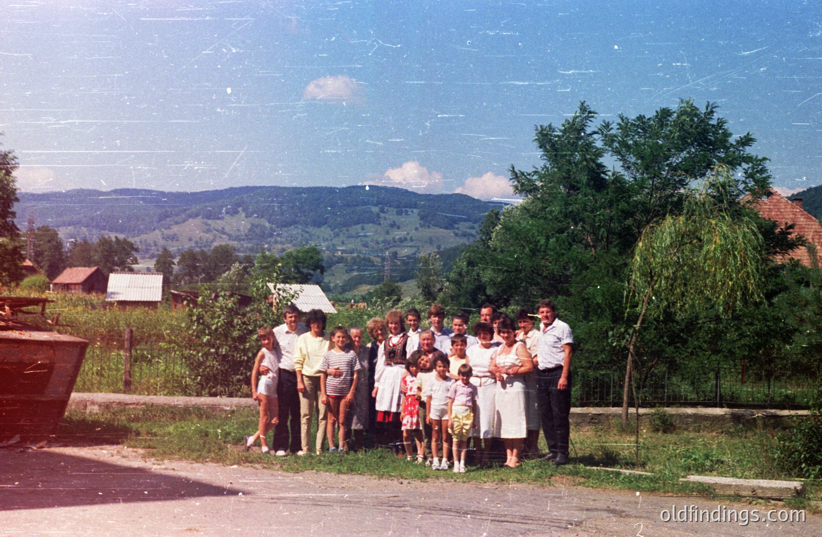 Group portrait of a large family posed outdoors before a backdrop of rolling hills and traditional rural buildings. Subjects wear 1970s-era clothing. Likely a community gathering in a rural European setting. A weathered look indicates age and potential archival value.