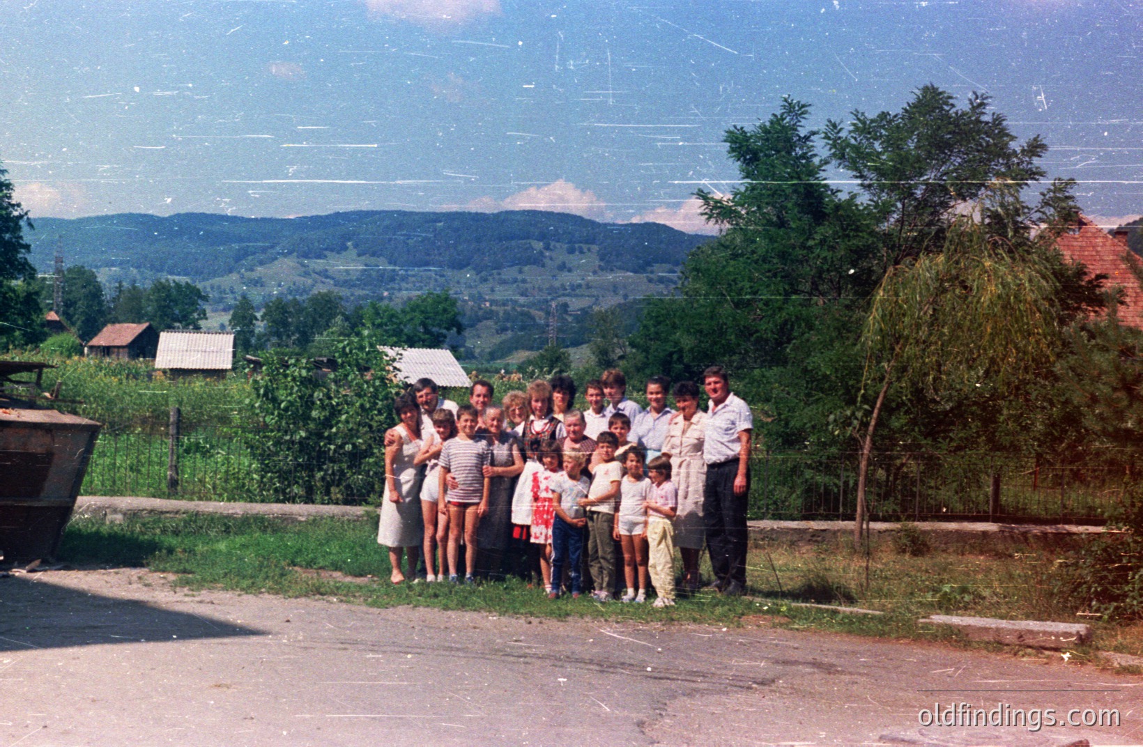 A group portrait features numerous children and several adults in front of rural homes against a backdrop of forested hills. Likely Eastern Europe, possibly Yugoslavia or Romania, 1970s based on clothing & photographic style. Simple architecture, overgrown vegetation, natural setting. Shows a community gathering.
