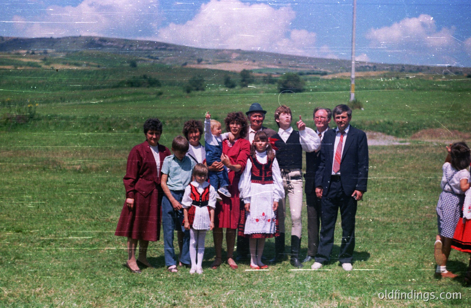A family group poses outdoors amidst a lush green meadow with distant hills visible. Several individuals wear traditional, folk-inspired clothing featuring red and black patterns. The group appears to be celebrating a cultural event or festival. Likely 1970s or 1980s.