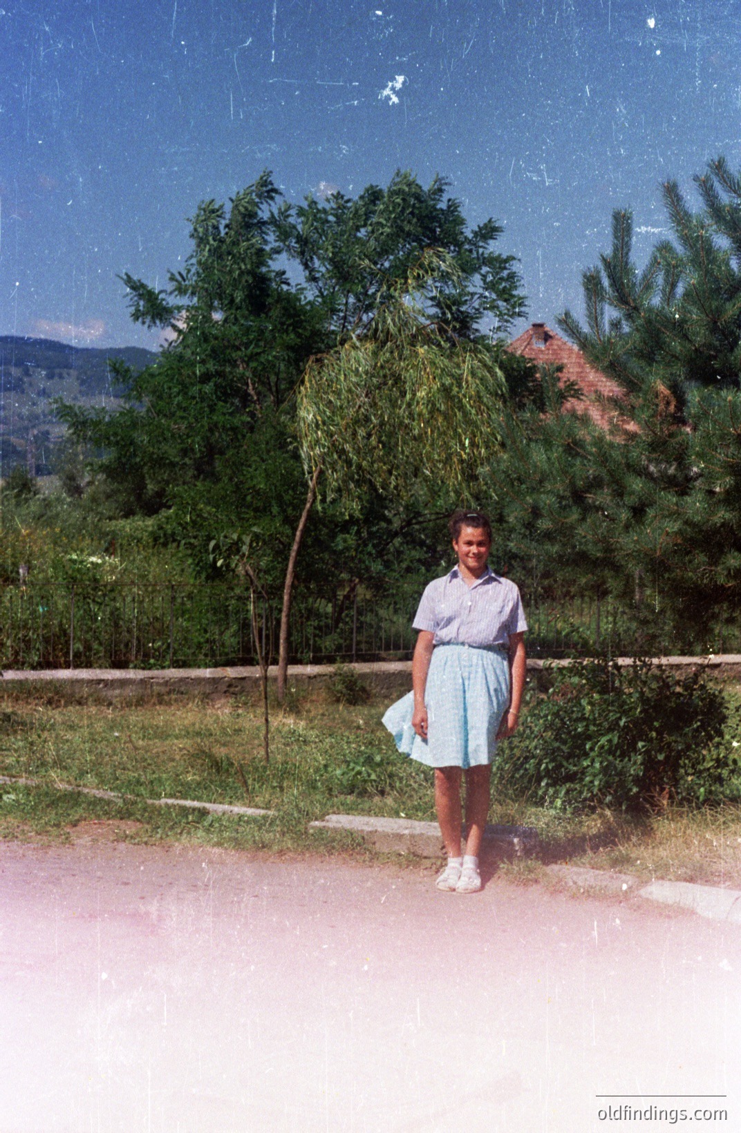 A young woman stands on a gravel path beside a willow tree, likely in Bulgaria. She wears a light blue, patterned dress and white shoes. A residential building is visible in the background amidst lush greenery and distant mountains. Appears to be a candid snapshot from the 1970s.