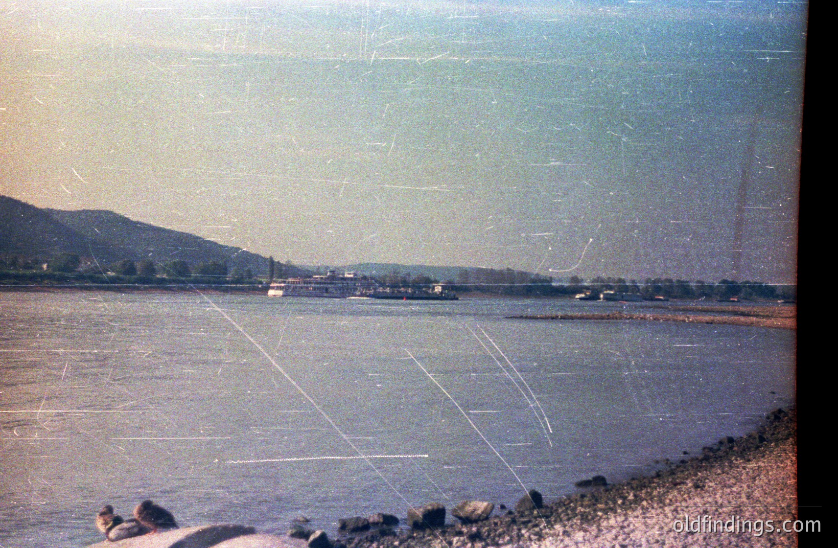 Seascape view with a river or bay, featuring a large passenger boat. Gentle slope with greenery and a rocky shoreline. Appears to be a vintage color print, exhibiting light scratches/wear. Possibly a resort or coastal town setting. Likely 1970s or 1980s.