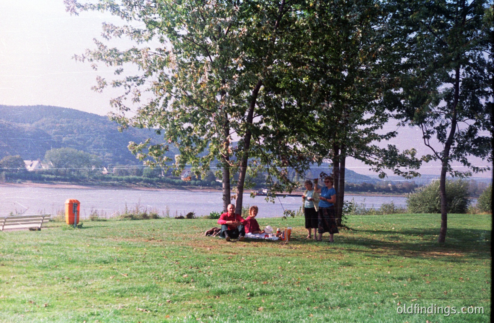 Family picnic scene: three people seated on a blanket beneath trees overlook a wide river with a bridge visible in the distance. Lush green lawn & hillside backdrop suggest a park or recreational area. Likely 1980s based on film quality & clothing styles. Stock photo potential for family leisure.