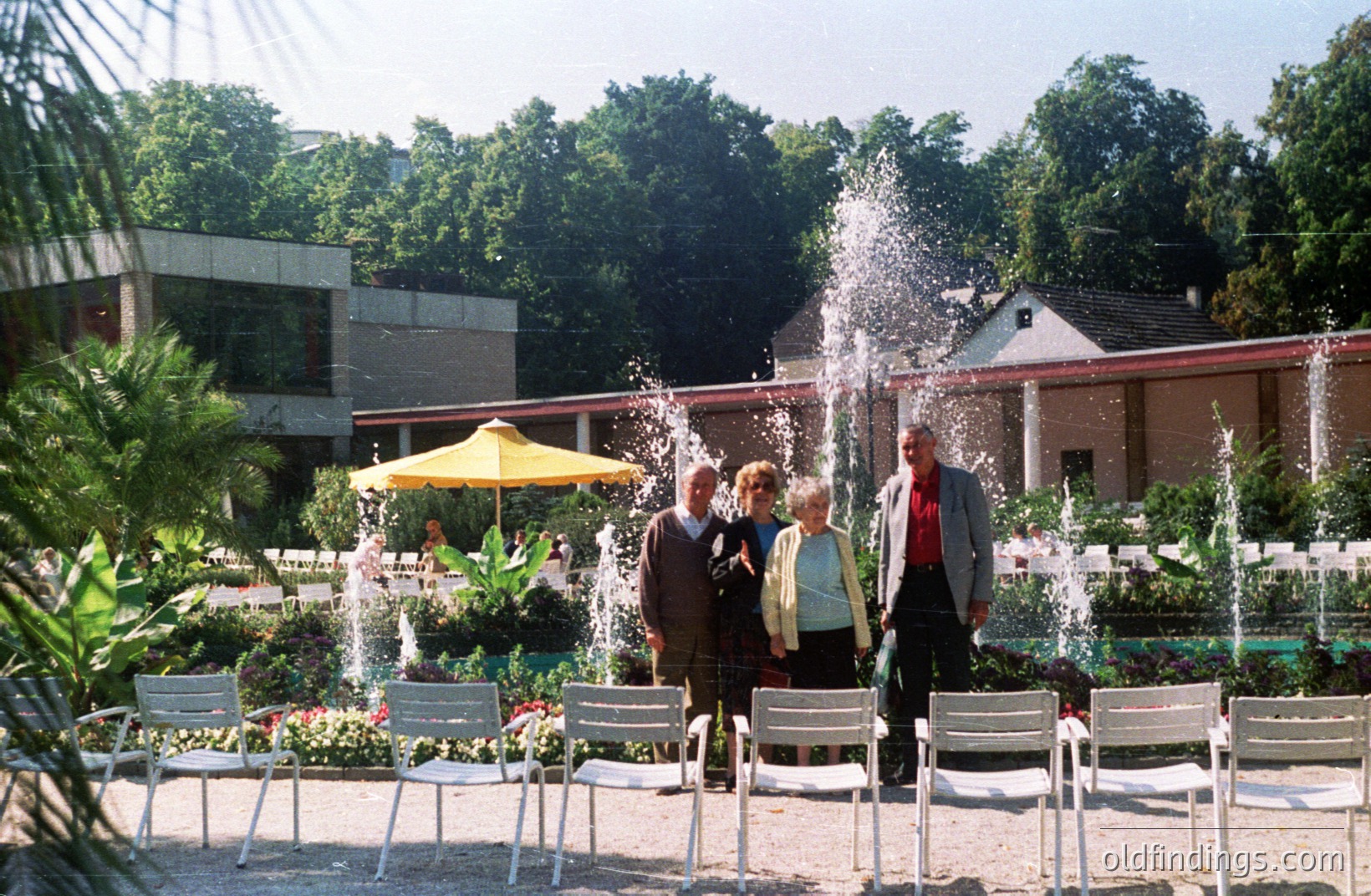 Group of formally-dressed individuals stands before tiered fountains and a mid-century modern building. Lush greenery and palm trees frame the scene. A yellow parasol adds a pop of color. Likely a public park or resort setting.