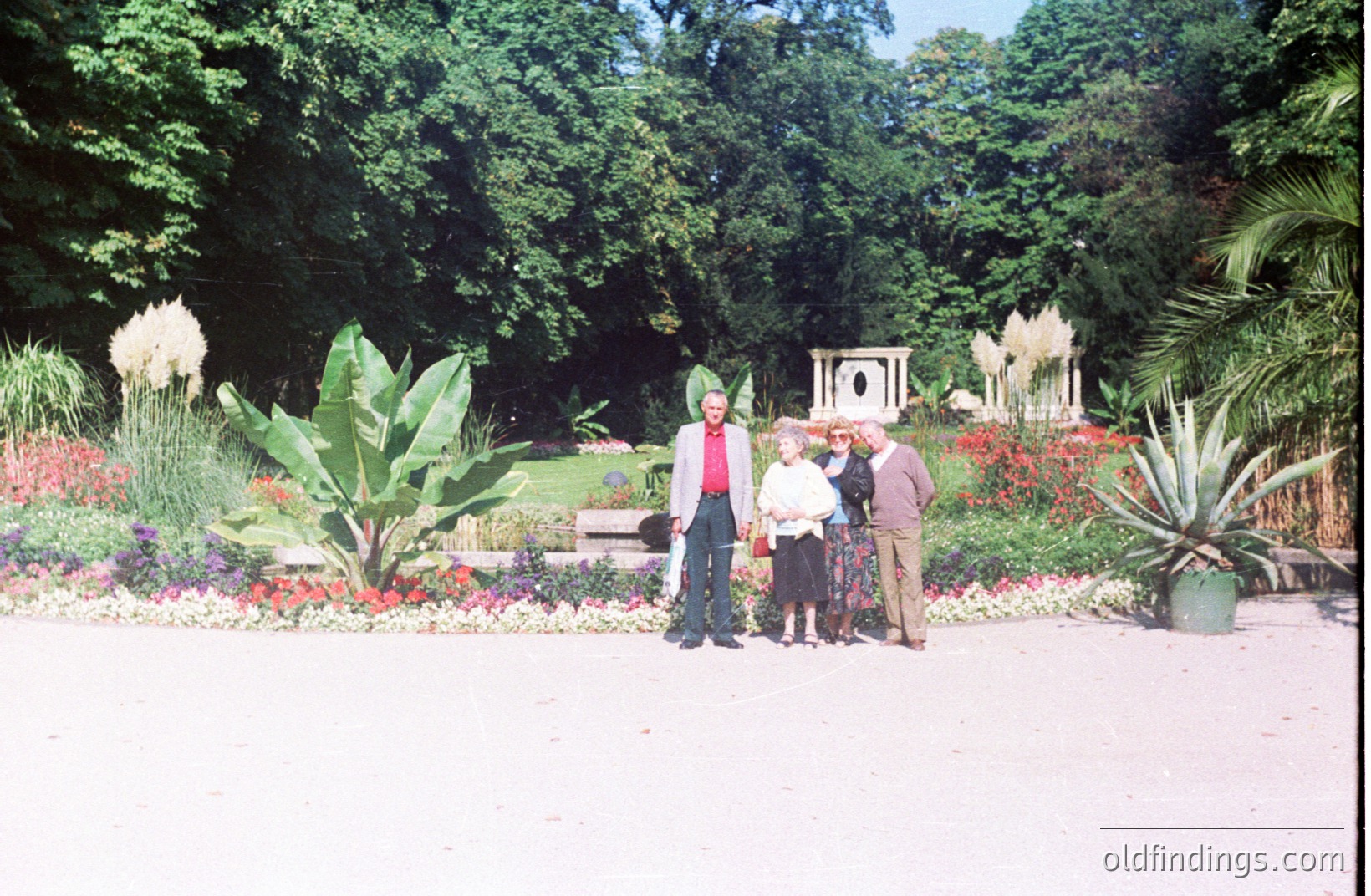 Four figures stand before a formal garden display featuring tiered flowerbeds, ornamental grasses, and a central stone structure. The group, appearing to be a family, is positioned on a gravel path within a lush, green environment. Likely a public park, potentially 1970s era.