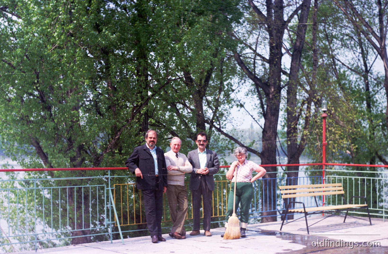 Four men pose near a green and yellow railing overlooking a body of water and lush foliage. Formal business attire contrasts with the casual attire of the fourth man, holding a broom. Likely a public park setting, possibly 1970s. Shows recreational space and community management.