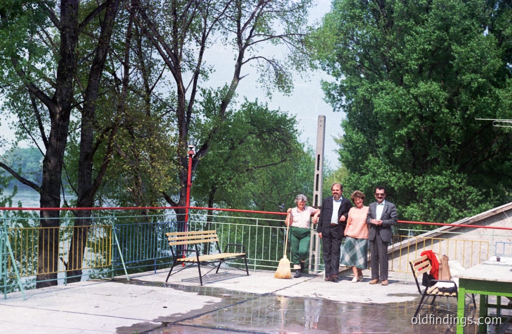 Outdoor scene: Four people stand on a patio overlooking a river, likely a resort or public space. Architecture includes a low, tiled roof and a decorative metal railing. The style hints at a 1970s aesthetic. Brooms and outdoor furniture are present.