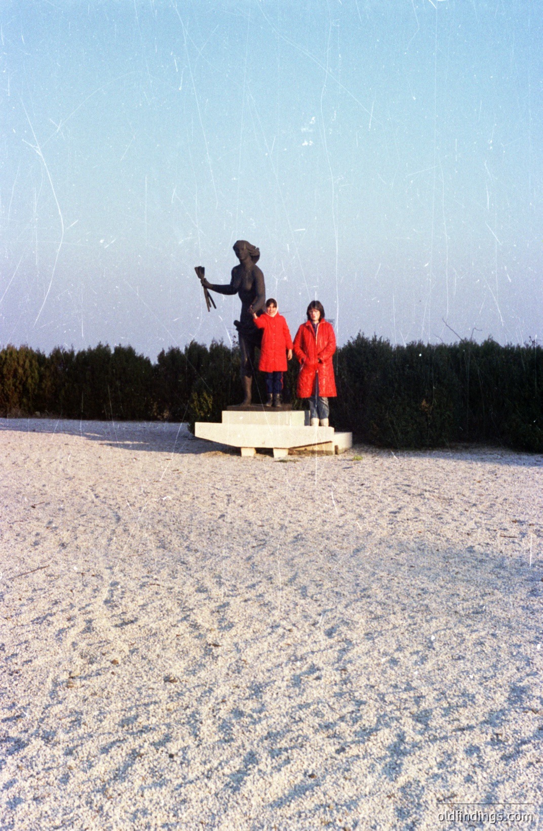 A bronze statue depicting a man in historical military attire stands on a stone pedestal. Two children in matching red parkas pose before it on a snow-covered expanse. Likely a commemorative monument; potentially a seaside location. The scene suggests a cold climate & mid-20th century aesthetic.