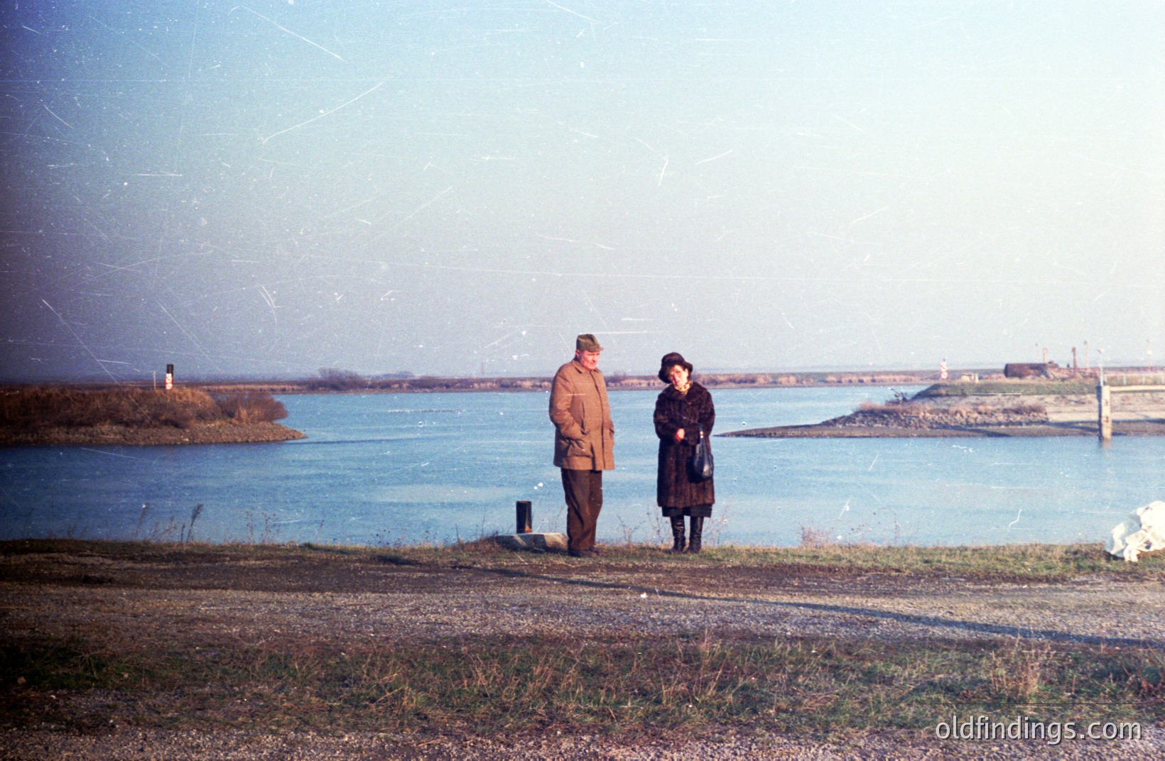 A color photograph depicts a couple standing on a grassy verge overlooking a wide body of water, possibly a bay or inlet. The pair are dressed in 1960s-70s style outerwear. A distant industrial complex is visible along the shoreline. Likely coastal Bulgaria, Varna region. Potential stock use for travel or fashion.