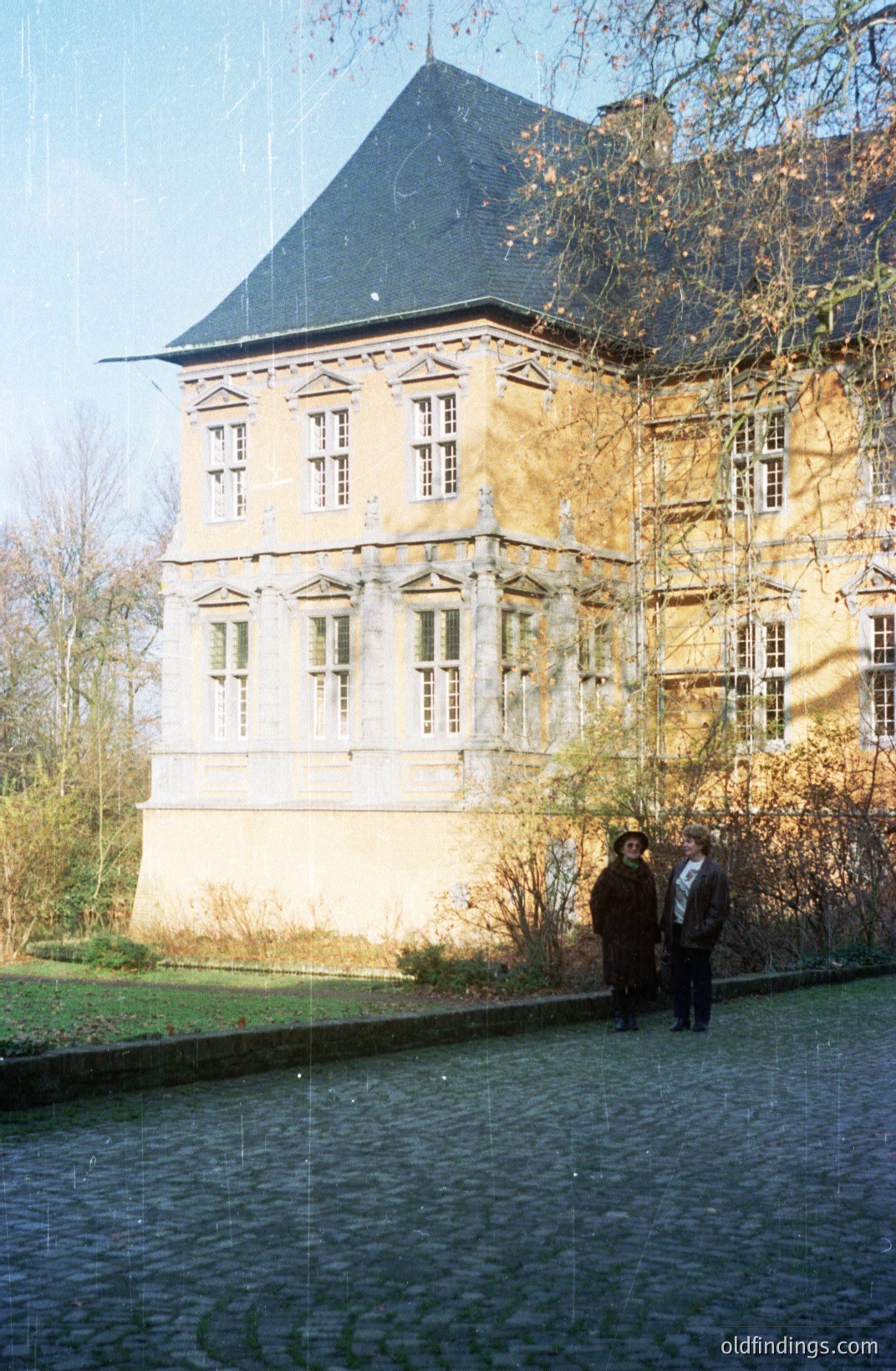 A stately, multi-story building with a dark slate roof and extensive scaffolding suggests ongoing restoration. The facade features many windows and decorative elements, partially obscured by the protective structures. Two figures in coats stand in the foreground near a reflective pool. Likely a European estate, possibly 20th-century.