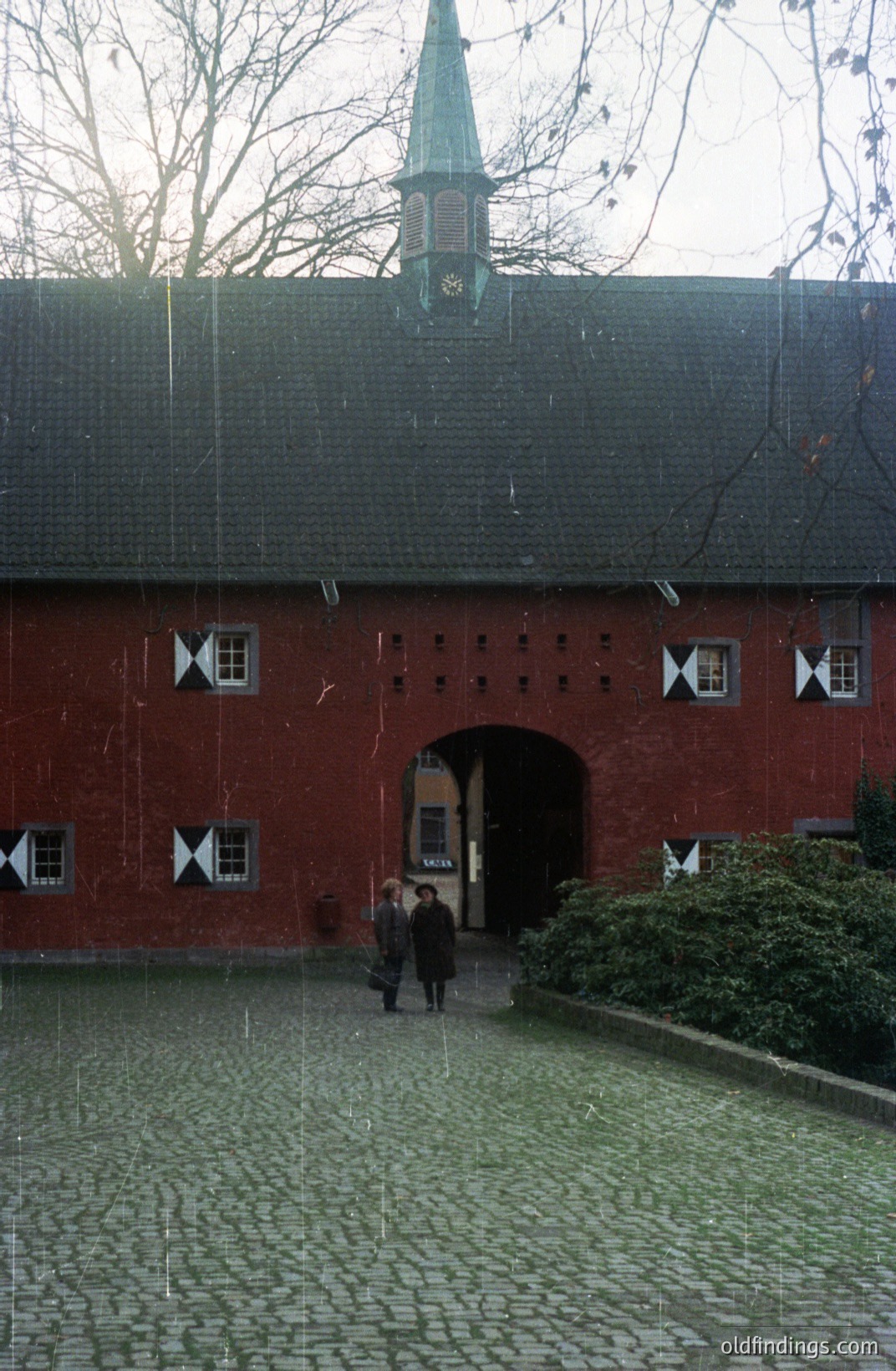 A symmetrical red brick building with decorative windows and a small tower. Two figures stand in a cobbled courtyard in light rain. The architecture suggests a European estate or castle. Likely mid-20th century, possibly 1960s-70s. Suitable for historical research or design reference.