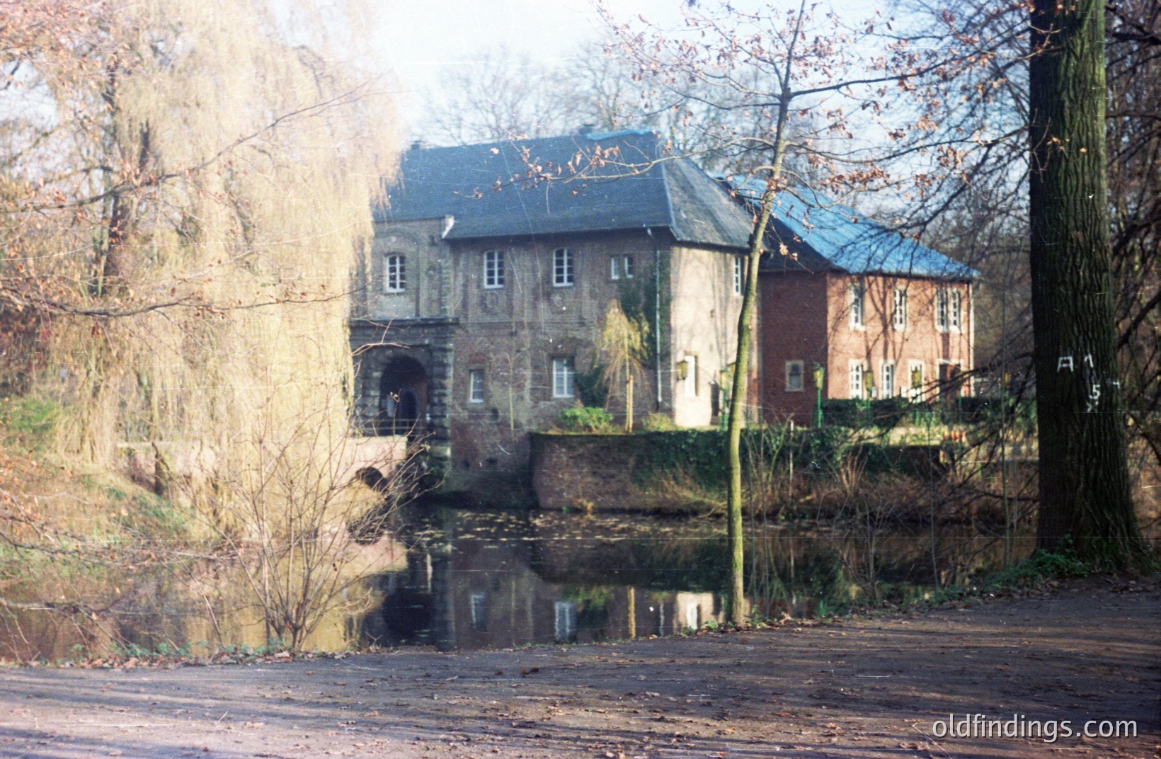 A historic mill building, constructed of brick and stone, stands beside a placid, reflective pond. The structure features multiple windows and a slate roof, with a visible water wheel entry. Appears to be a rural or semi-rural setting. Likely 20th century, possibly earlier.